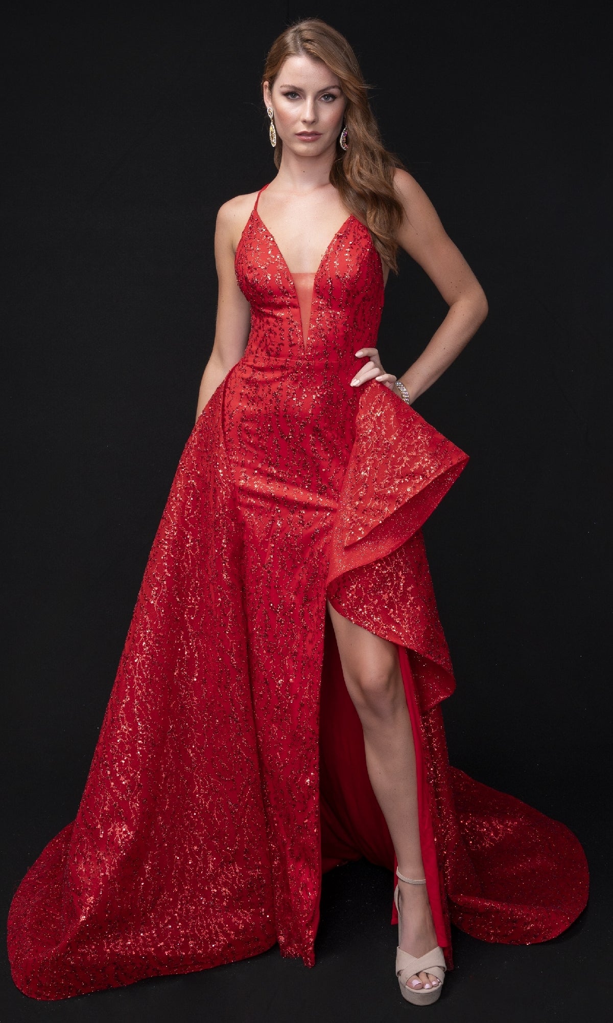 A woman poses against a black background wearing the Long Formal Dress: Nina Canacci 8250, a sleeveless sparkling red gown with a thigh-high slit and ruffled overskirt, accessorized with earrings and platform heels.